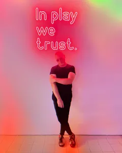 Standing under a neon sign reading “In play we trust”.